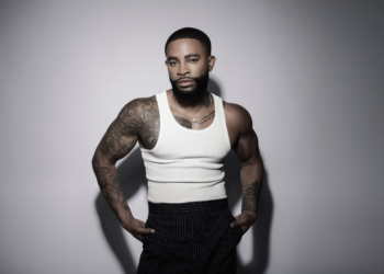 A photo of singer Elijah Blake wearing a white tank top, black slacks, against a gray backdrop