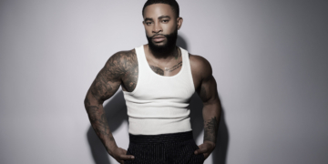 A photo of singer Elijah Blake wearing a white tank top, black slacks, against a gray backdrop