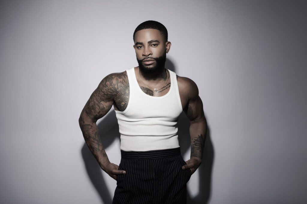 A photo of singer Elijah Blake wearing a white tank top, black slacks, against a gray backdrop